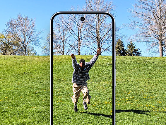 A man joyfully jumps in a grassy park, viewed through the frame of a Pixel phone
